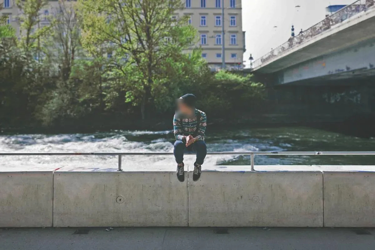 Adolescente con gorro sentado sobre un muro de hormigón junto a un río, con árboles y un puente al fondo
