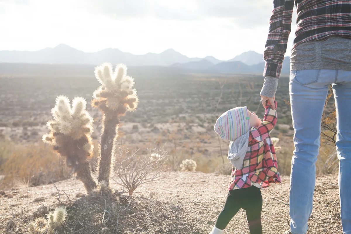 Adulto con camisa de cuadros y vaqueros da la mano a un niño pequeño con gorro de lana durante un paseo por un paisaje seco y soleado con montañas al fondo