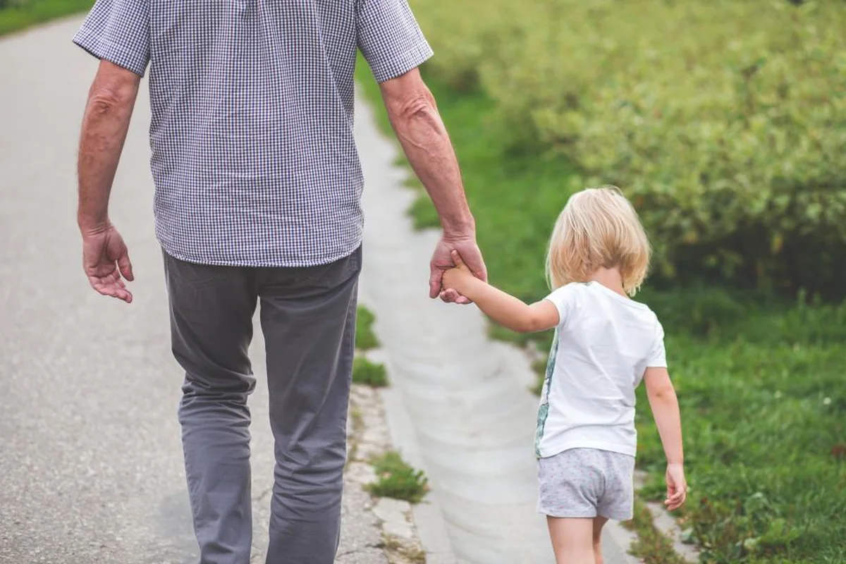 Un adulto y una niña pequeña caminan de espaldas por un camino junto a la hierba, cogidos de la mano