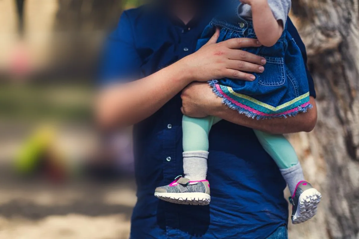 Un adulto con camisa azul lleva en brazos a una niña pequeña con falda vaquera y zapatillas, en un parque