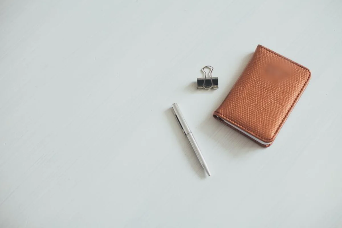 Una pequeña agenda de piel color cobre junto a un bolígrafo plateado y una pinza metálica sobre una mesa blanca