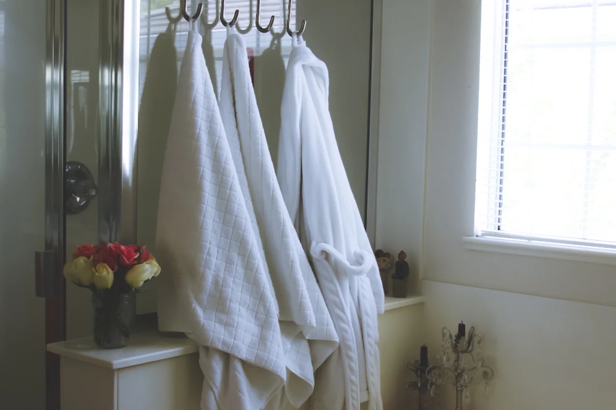 Tres albornoces blancos colgados de unos ganchos en un baño luminoso, con un jarrón de flores rojas y amarillas y una ventana con luz natural