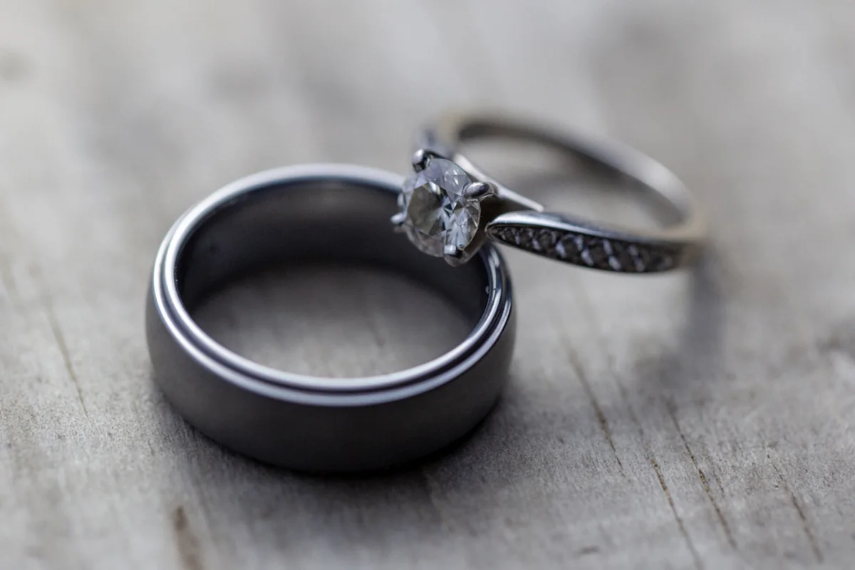 Alianza de metal oscuro y anillo de compromiso con un diamante apoyados uno junto al otro sobre una superficie de madera clara