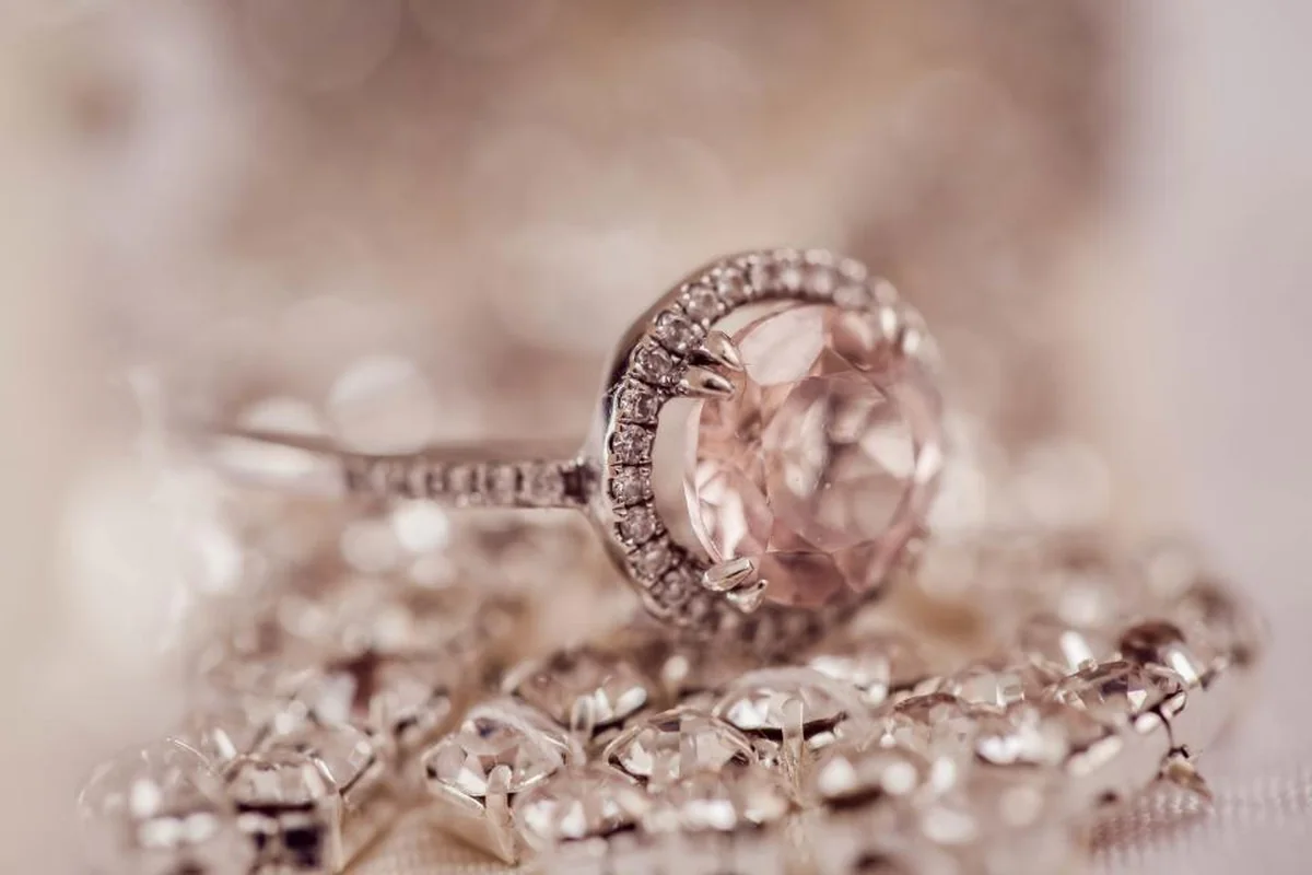 Anillo con una piedra rosada y pequeños brillantes apoyado sobre una cadena de cristales, con el fondo desenfocado en tonos rosados