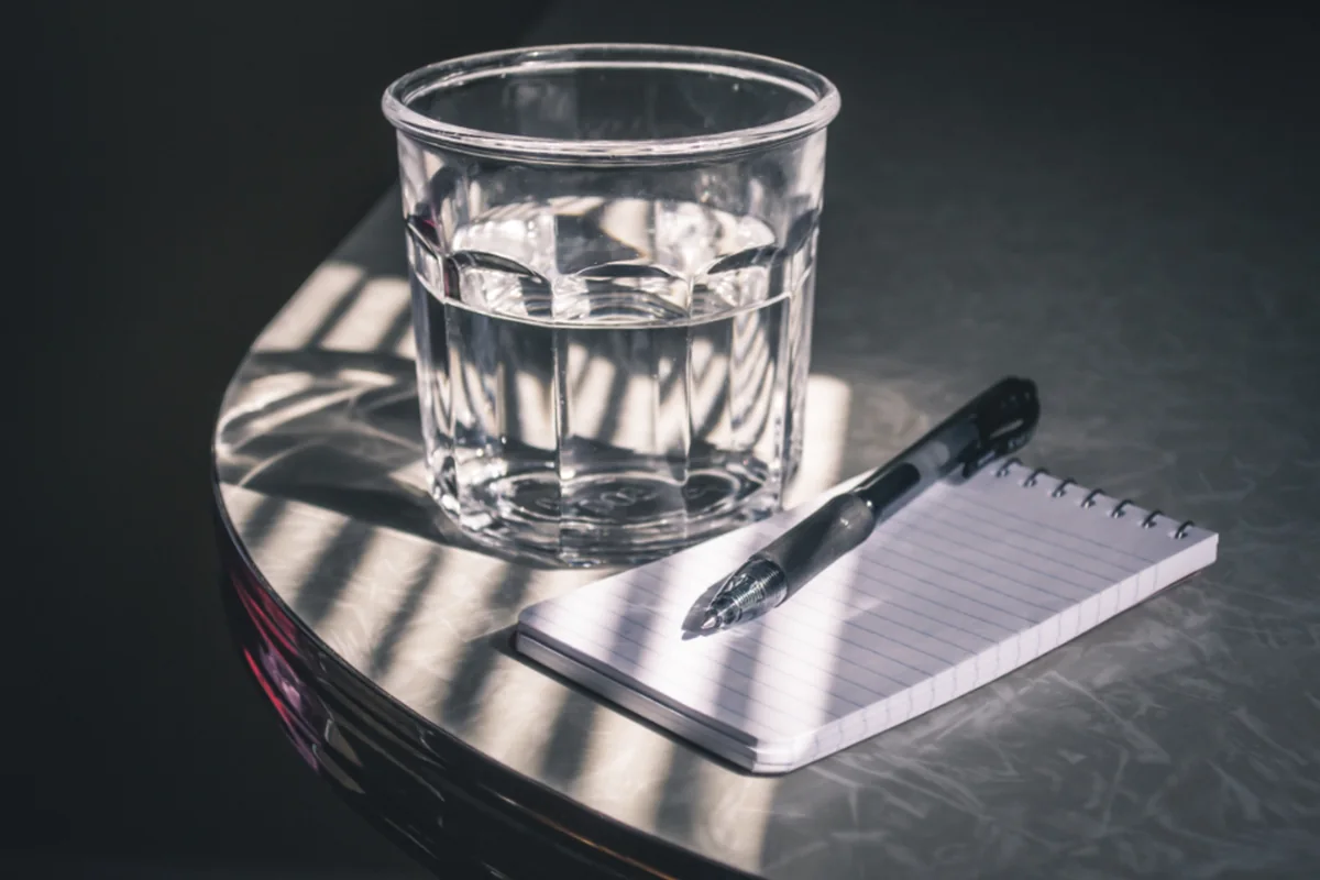 Libreta, bolígrafo y un vaso de agua sobre una mesa en penumbra