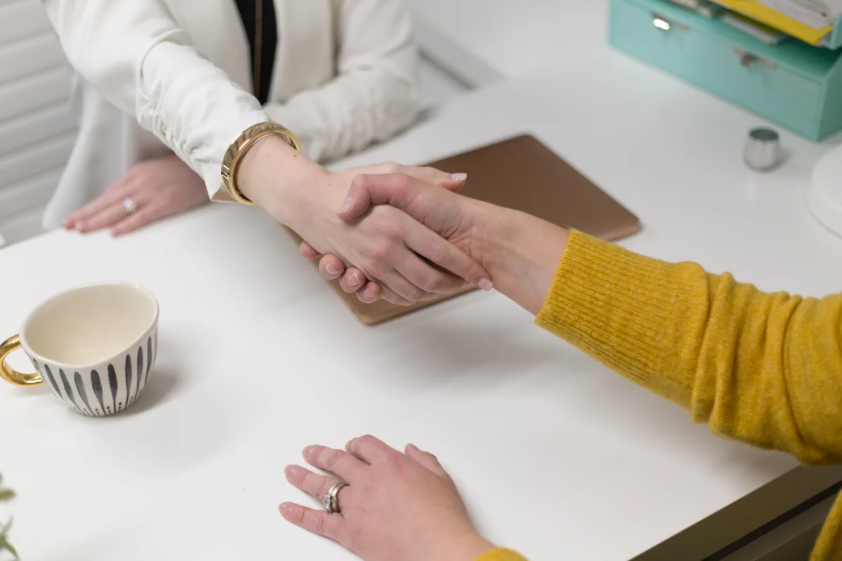 Dos personas se dan la mano sobre una mesa blanca de oficina con una taza de café y una carpeta