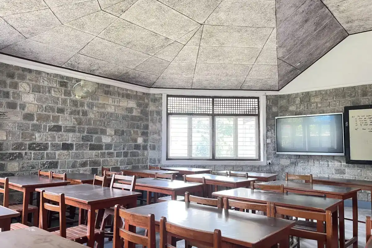 Aula vacía con mesas y sillas de madera, paredes de piedra, una ventana y una pantalla apagada en la pared