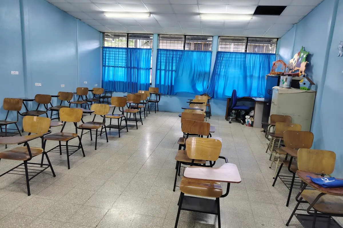 Aula vacía con filas de pupitres de madera, paredes azules y ventanales con cortinas