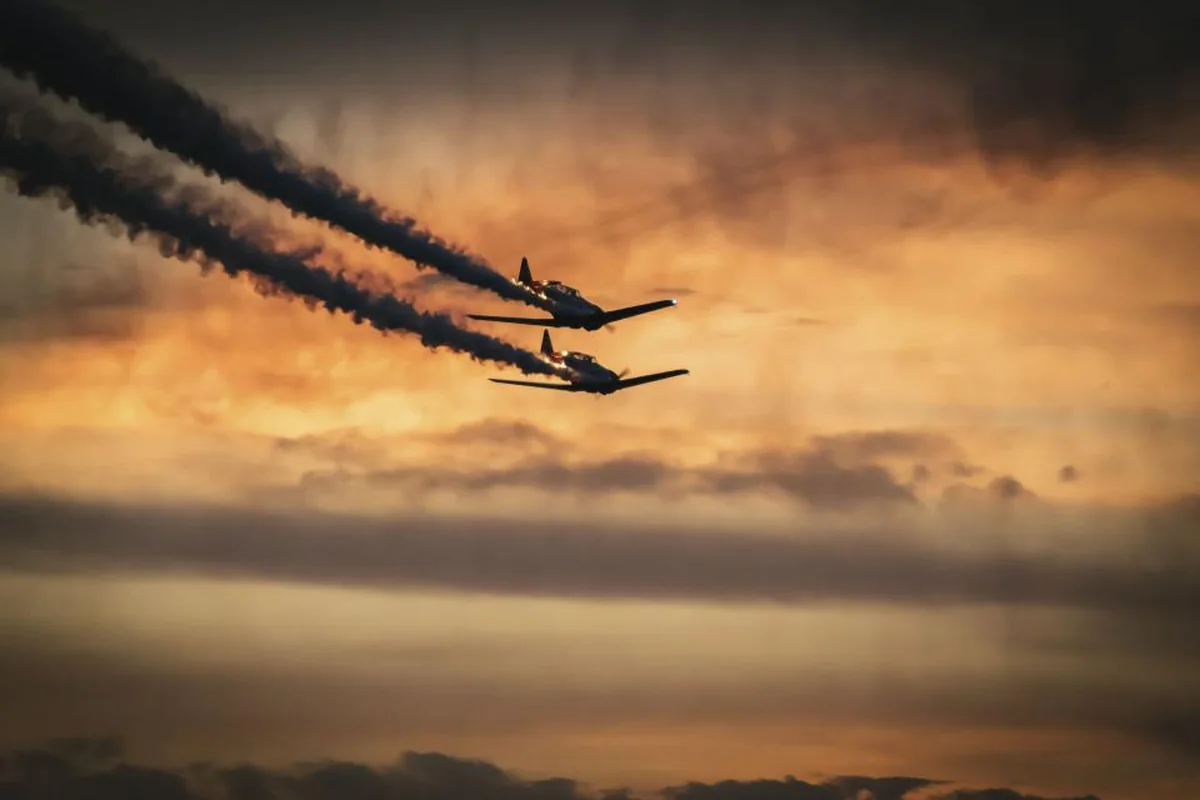 Dos avionetas en vuelo dejando una estela de humo oscuro sobre un cielo anaranjado al atardecer