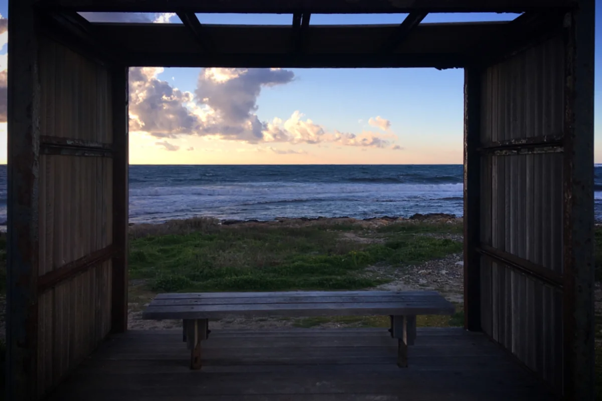 Un banco de madera vacío bajo un porche de tablones abierto al mar, con el cielo del atardecer al fondo