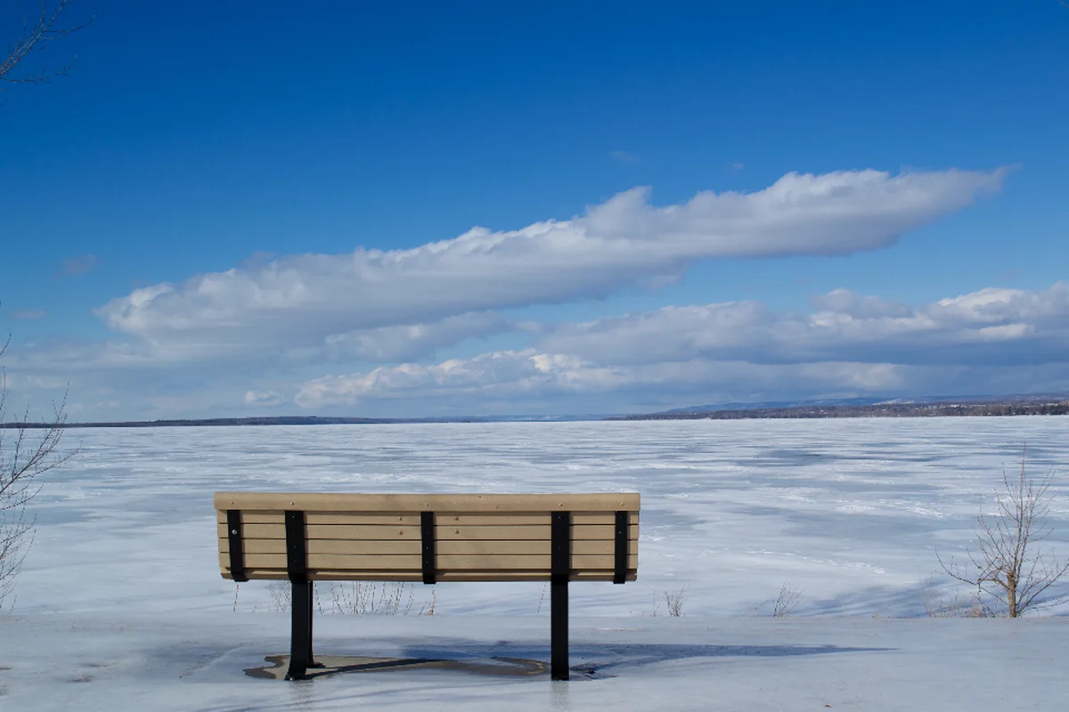 Banco de madera vacío frente a un lago helado bajo un cielo azul con nubes