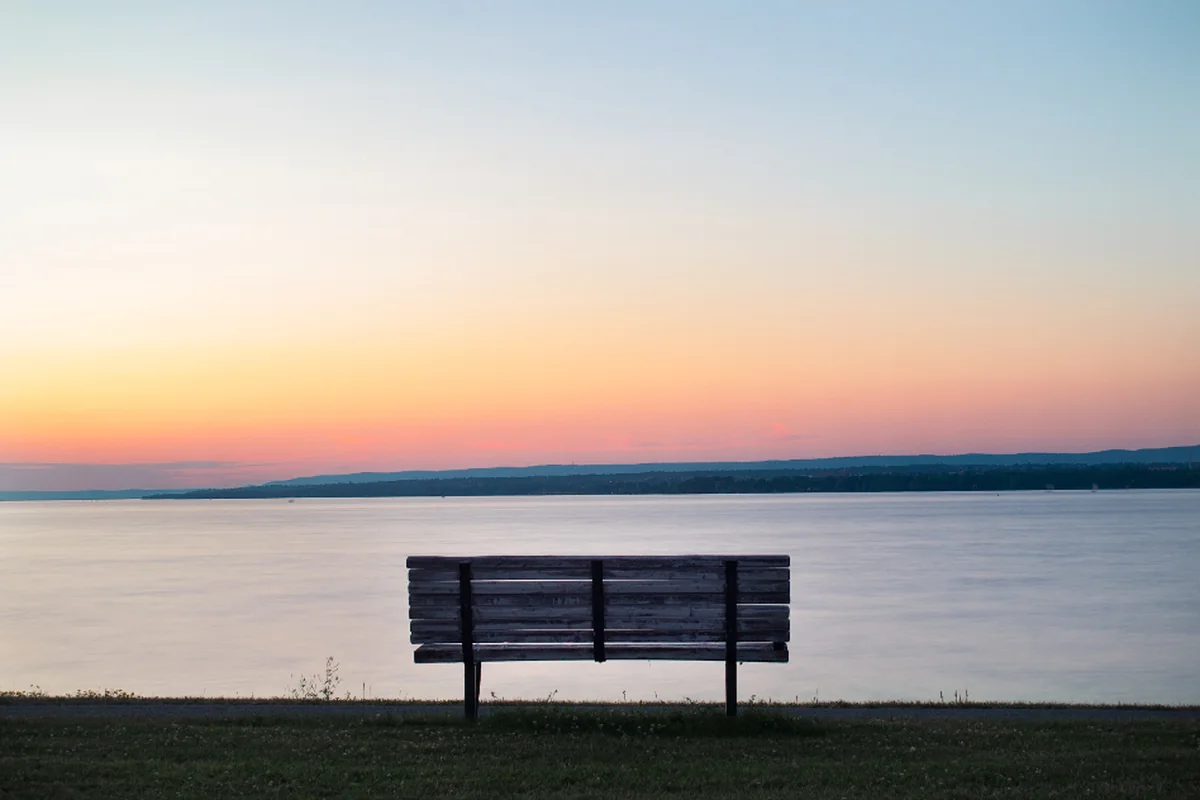 Banco de madera vacío frente a un lago en calma al atardecer, con el cielo anaranjado sobre la orilla