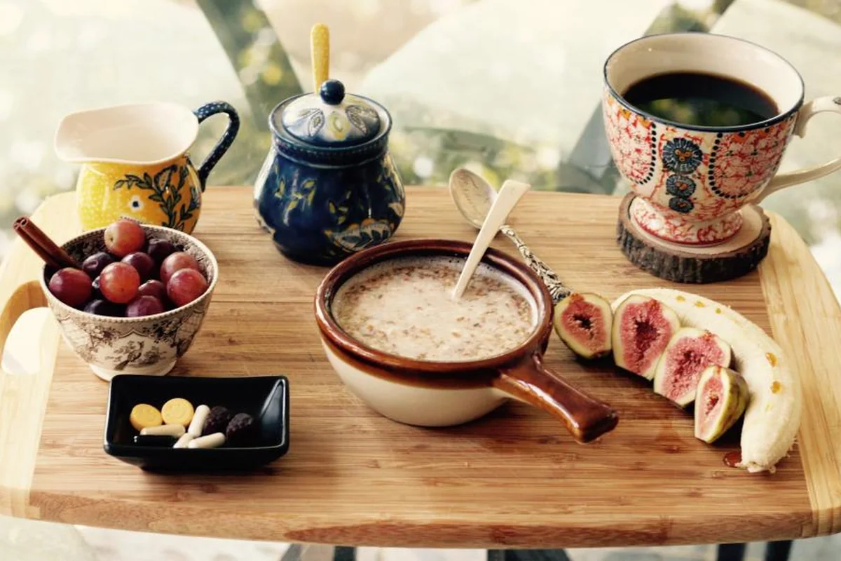 Bandeja de madera con un cuenco de uvas, un cuenco de gachas, higos abiertos, un plátano, una jarrita, un azucarero y una taza de café sobre una mesa de cristal