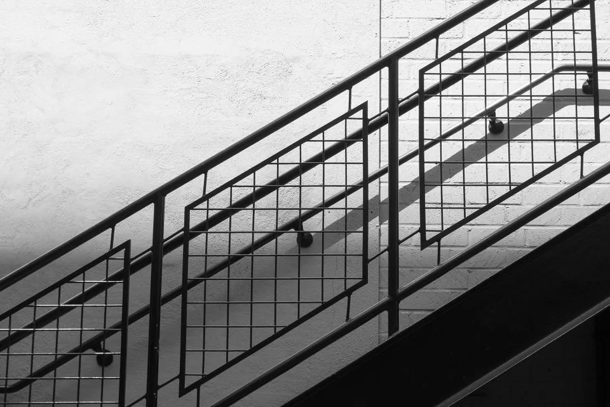 Barandilla metálica de una escalera en diagonal sobre una pared blanca, en blanco y negro