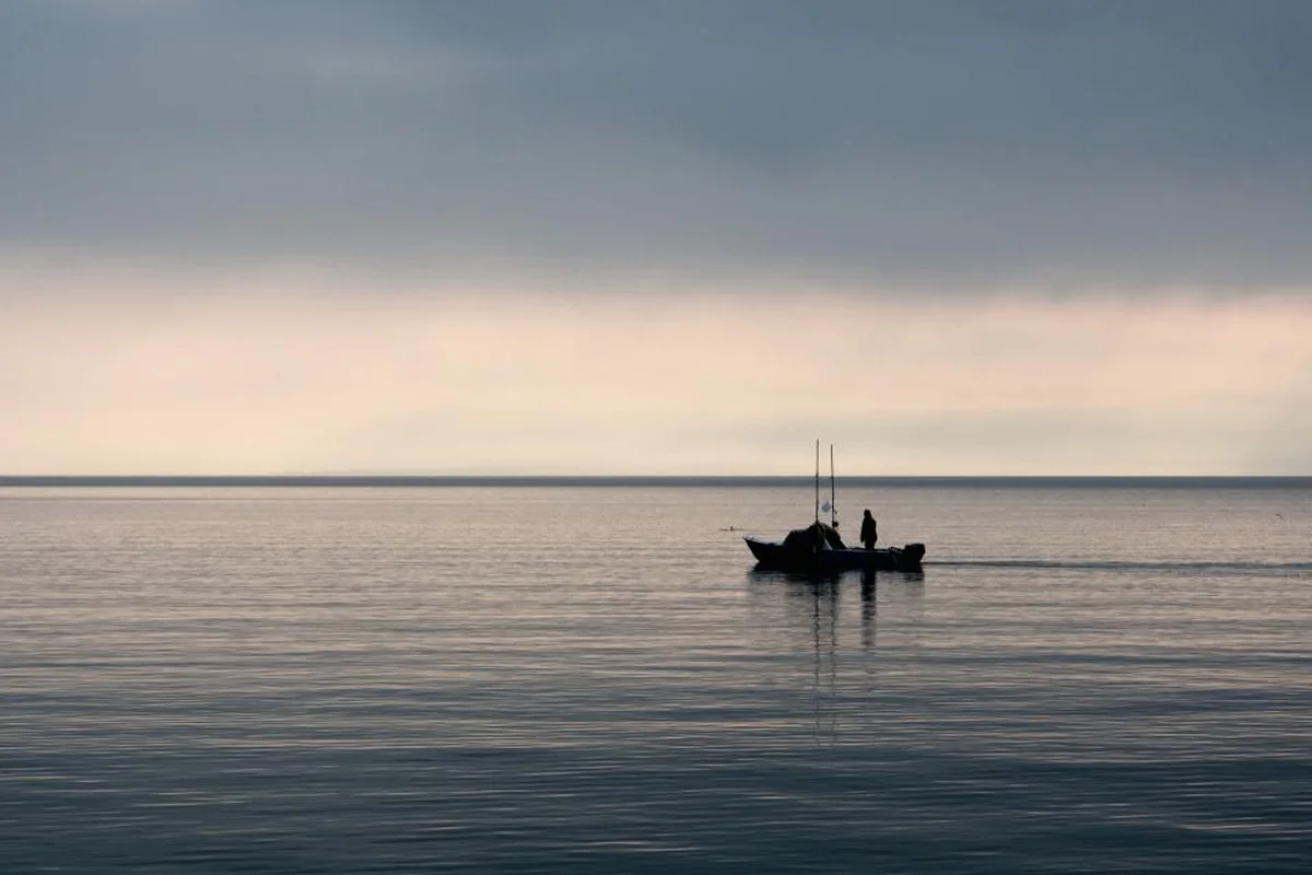 Una pequeña embarcación de pesca navegando sola en un mar en calma bajo un cielo nublado al atardecer, con el horizonte despejado
