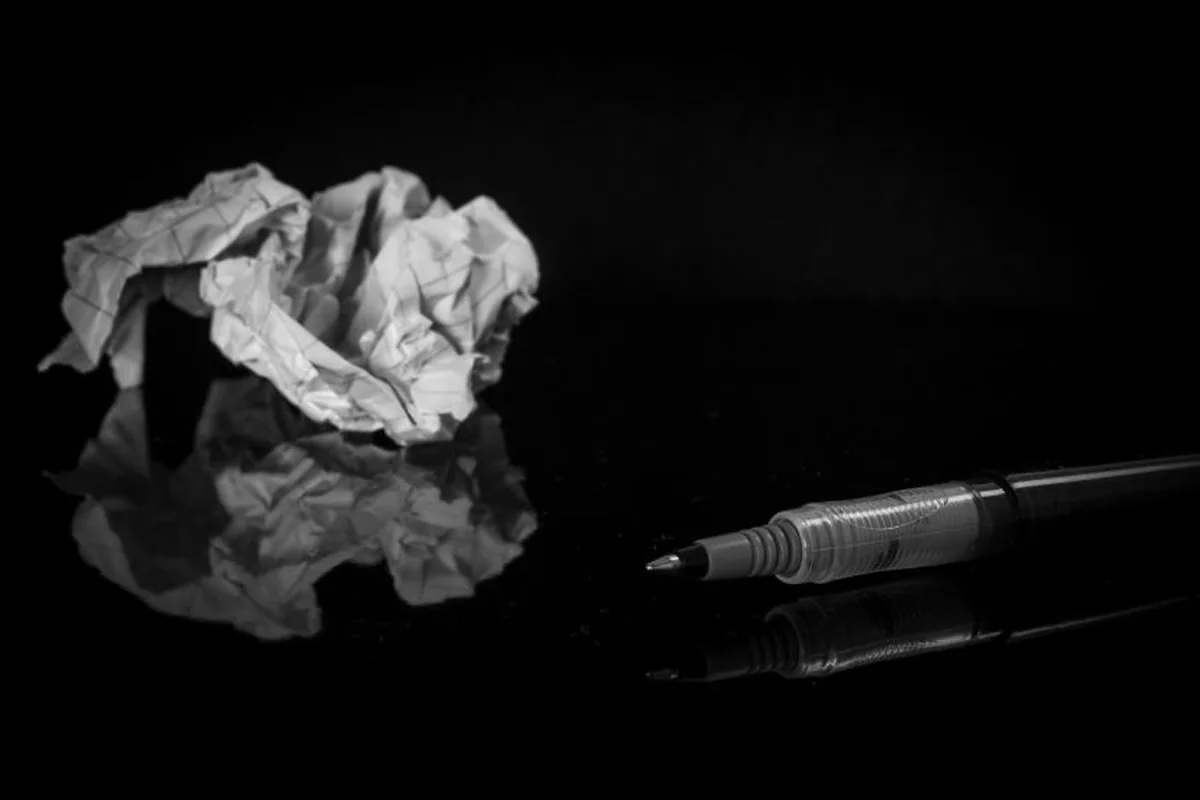 Una bola de papel arrugado y un bolígrafo sobre una superficie negra brillante que los refleja