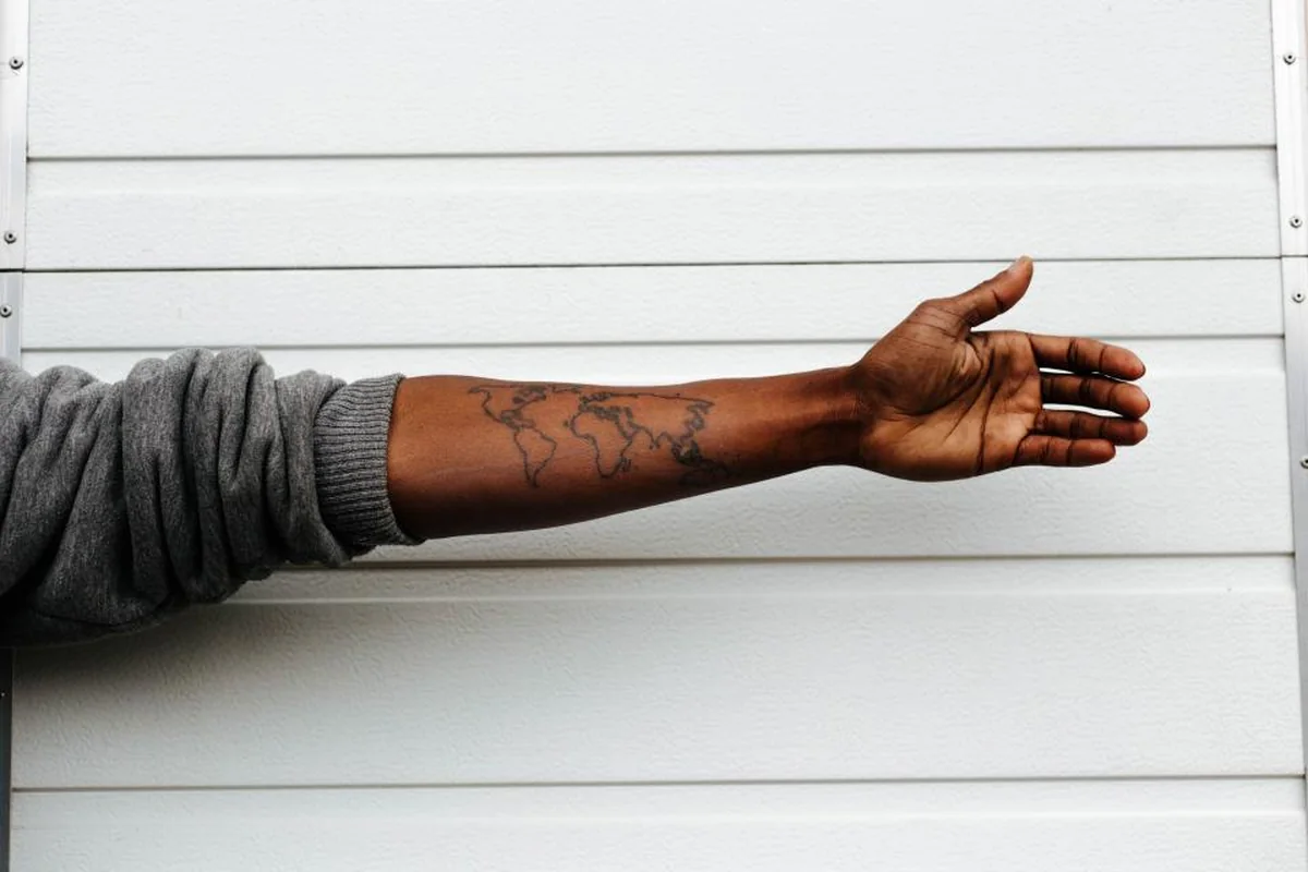 Antebrazo extendido con un tatuaje de un mapa del mundo en tinta negra, delante de una pared blanca de lamas