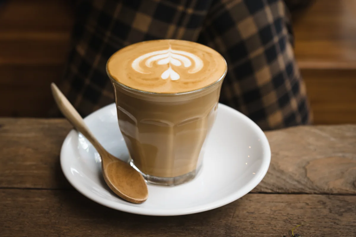 Café con leche con dibujo de espuma en un vaso de cristal sobre un plato blanco con cucharilla de madera, en una mesa de madera