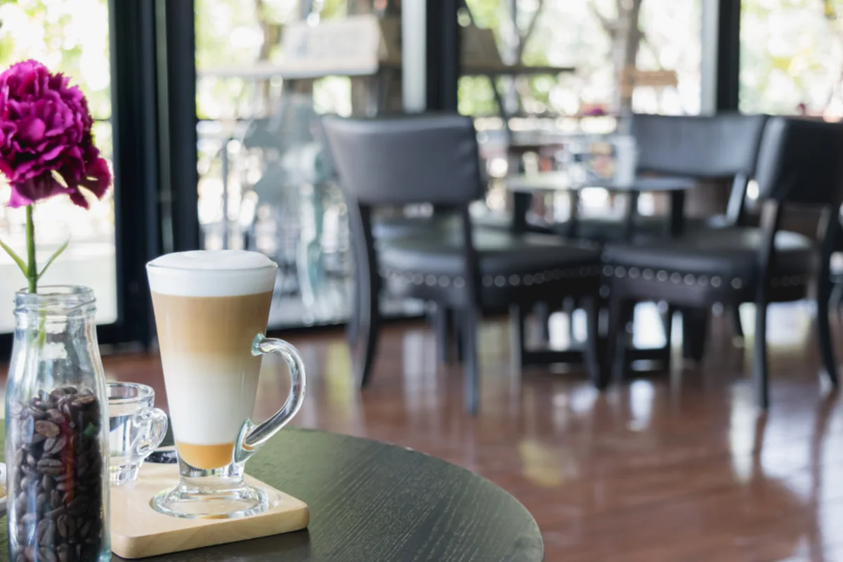 Vaso de café con leche sobre una mesa redonda de cafetería junto a un clavel en un tarro, con sillas vacías desenfocadas al fondo