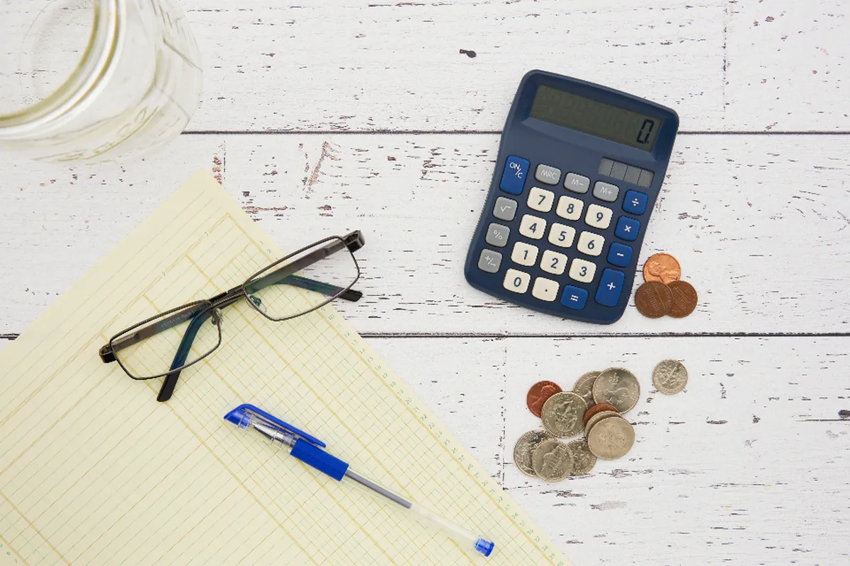 Calculadora azul, unas gafas, un bolígrafo, monedas sueltas y una hoja cuadriculada sobre una mesa de madera blanca