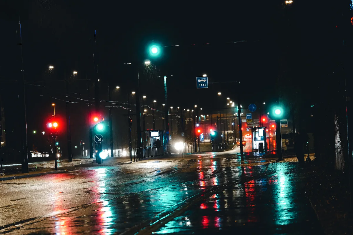 Calle mojada por la lluvia de noche, con semáforos verdes y rojos encendidos reflejados en el asfalto y farolas a lo lejos