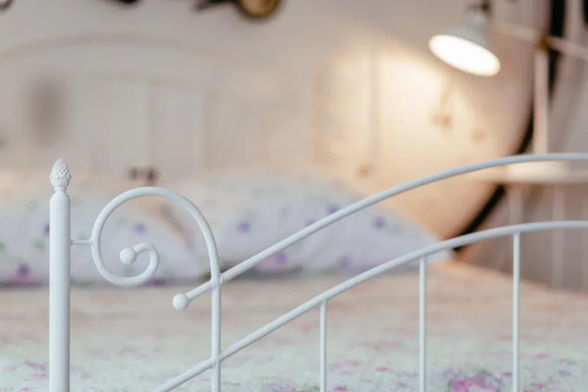 Pie de una cama de forja blanca en un dormitorio con luz cálida y almohadas de flores