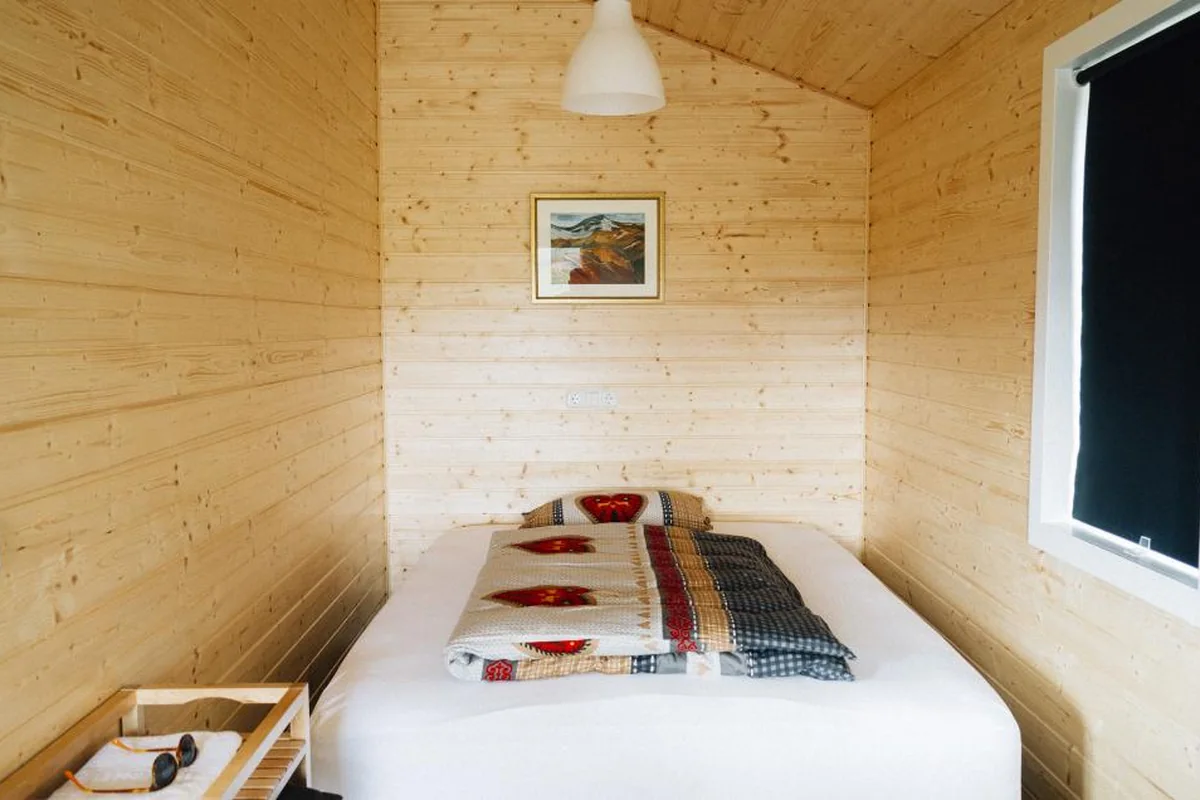Una cama con edredón doblado en una pequeña habitación de madera con una lámpara blanca y un cuadro en la pared