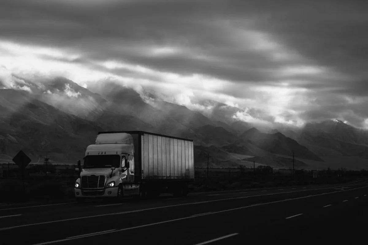 Camión con remolque circulando por una carretera bajo un cielo nublado y montañas, en blanco y negro