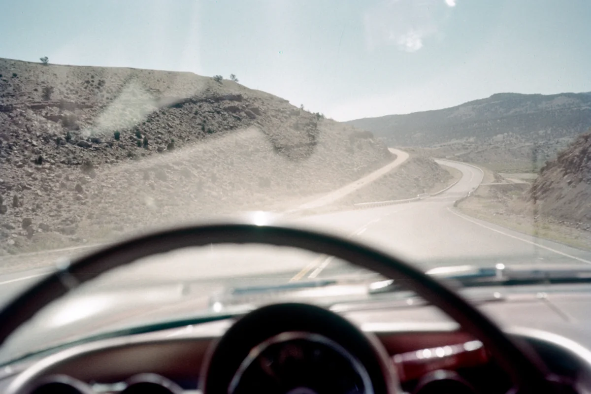 Carretera con curvas entre montañas áridas vista a través del parabrisas de un coche antiguo, con el volante en primer plano