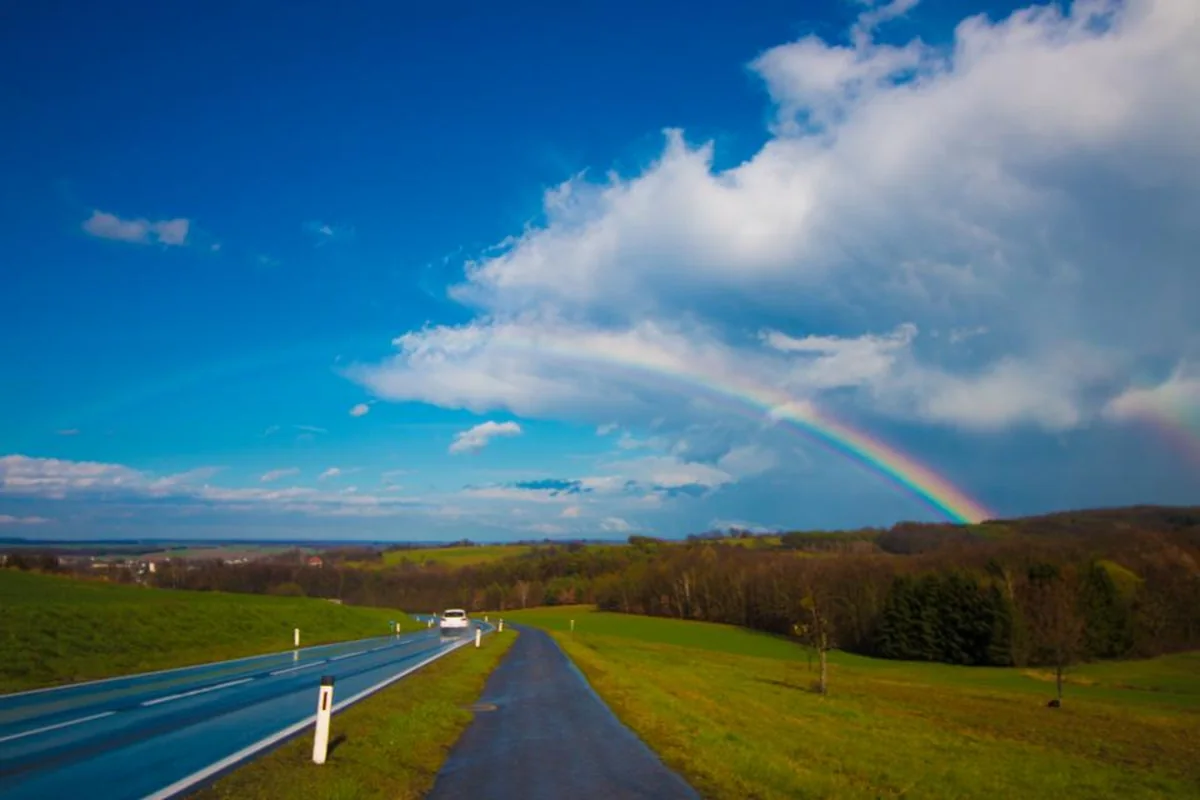 Carretera asfaltada y camino estrecho que se separan entre campos verdes bajo un cielo azul con un arcoíris