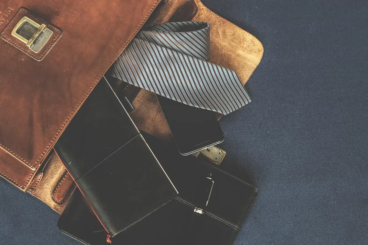 Cartera de piel marrón entreabierta con una corbata de rayas, un móvil y dos carteras negras asomando sobre una tela azul oscura