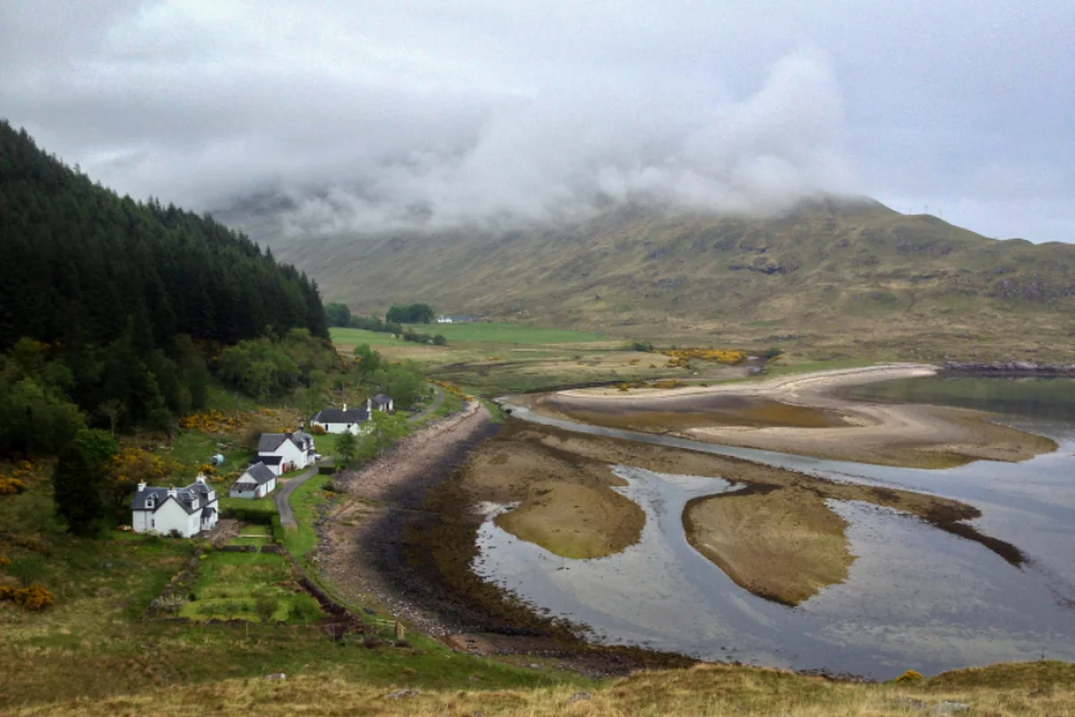 Unas pocas casas blancas aisladas junto a un río en un valle verde con montañas cubiertas de niebla
