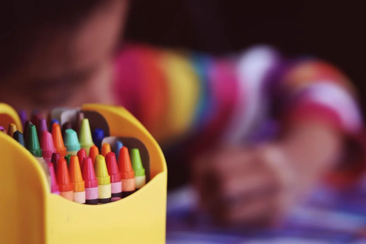 Bote amarillo lleno de ceras de colores en primer plano y, al fondo y muy desenfocado, un niño con jersey de rayas dibujando