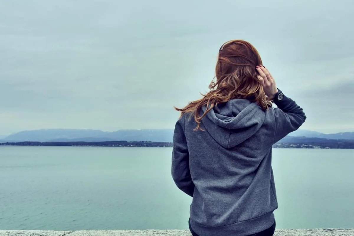Una joven con sudadera gris, de espaldas, sentada frente a un lago con montañas al fondo en un día nublado