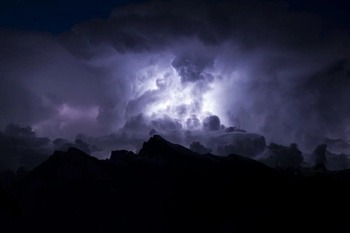 Un cielo nocturno cargado de nubes de tormenta iluminadas por un relámpago sobre la silueta oscura de unas montañas