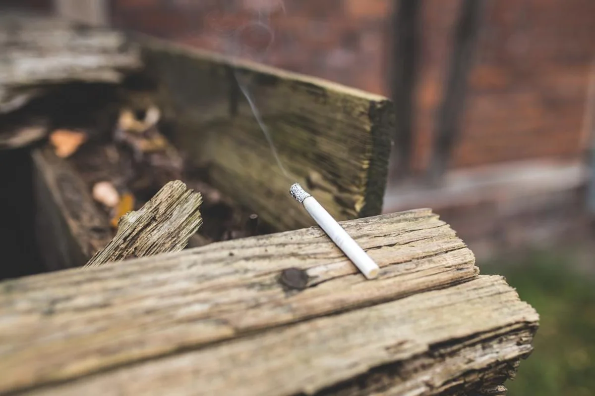 Cigarrillo encendido apoyado sobre una tabla de madera vieja, con un hilo de humo y el fondo desenfocado