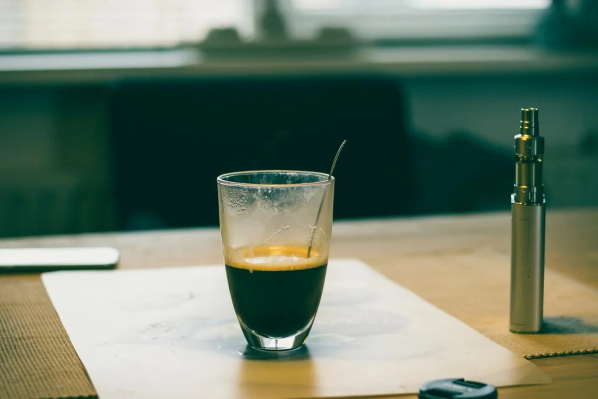 Cigarrillo electrónico metálico de pie junto a un vaso de café sobre una mesa de madera