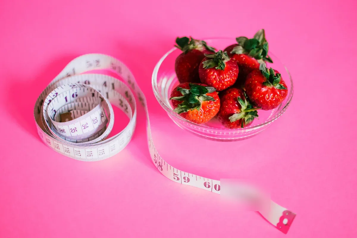 Una cinta métrica enrollada junto a un cuenco de cristal con fresas sobre un fondo rosa liso