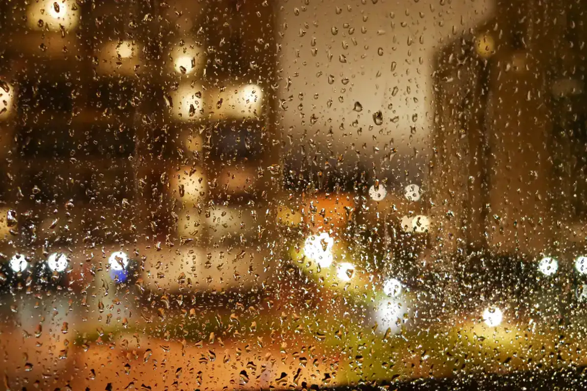 Luces cálidas de una ciudad de noche, desenfocadas tras un cristal cubierto de gotas de lluvia