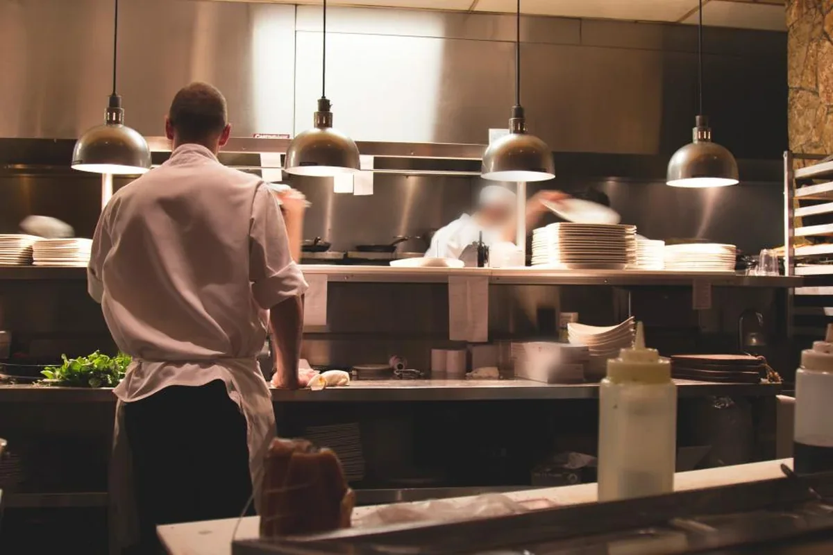 Cocinero de espaldas con chaquetilla blanca trabajando en la cocina de un restaurante bajo lámparas colgantes, con pilas de platos al fondo