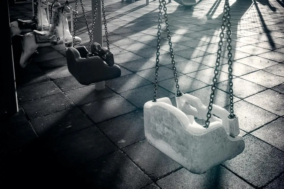Columpios infantiles de asiento cerrado vacíos en un parque, fotografiados en blanco y negro con sombras alargadas sobre el suelo
