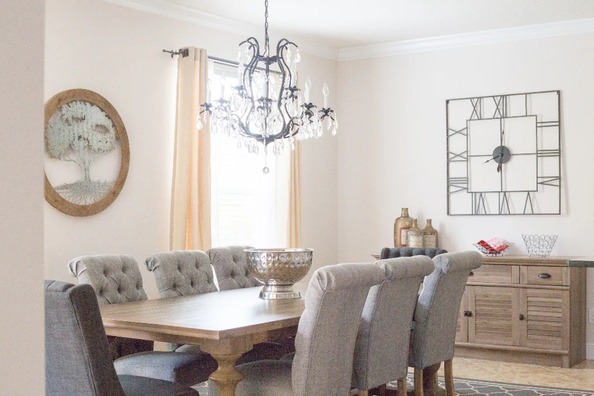 Comedor luminoso con una mesa de madera, sillas tapizadas en gris, una lámpara de araña y un reloj grande en la pared