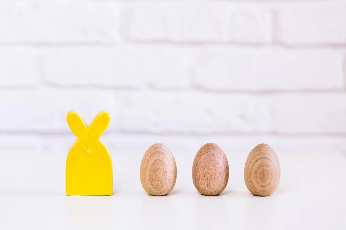 Conejito amarillo de madera junto a tres huevos de madera alineados sobre una mesa blanca