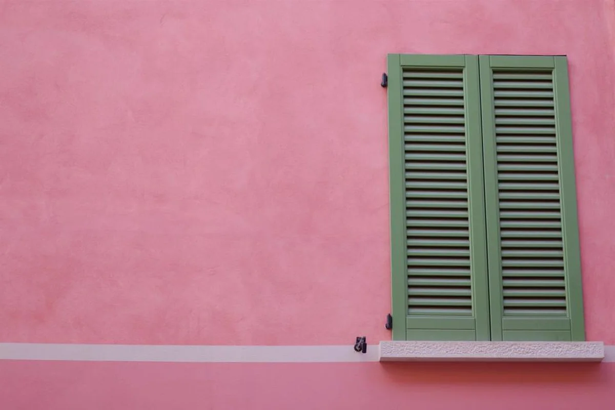 Contraventanas verdes de madera cerradas en una fachada lisa pintada de rosa, con un alféizar blanco