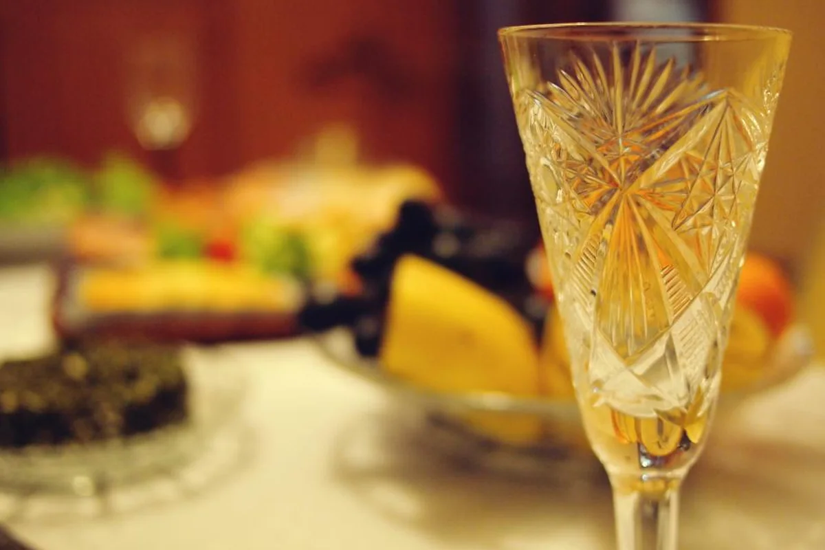 Copa de cristal labrado en primer plano sobre una mesa con mantel blanco, con fuentes de fruta y comida desenfocadas al fondo