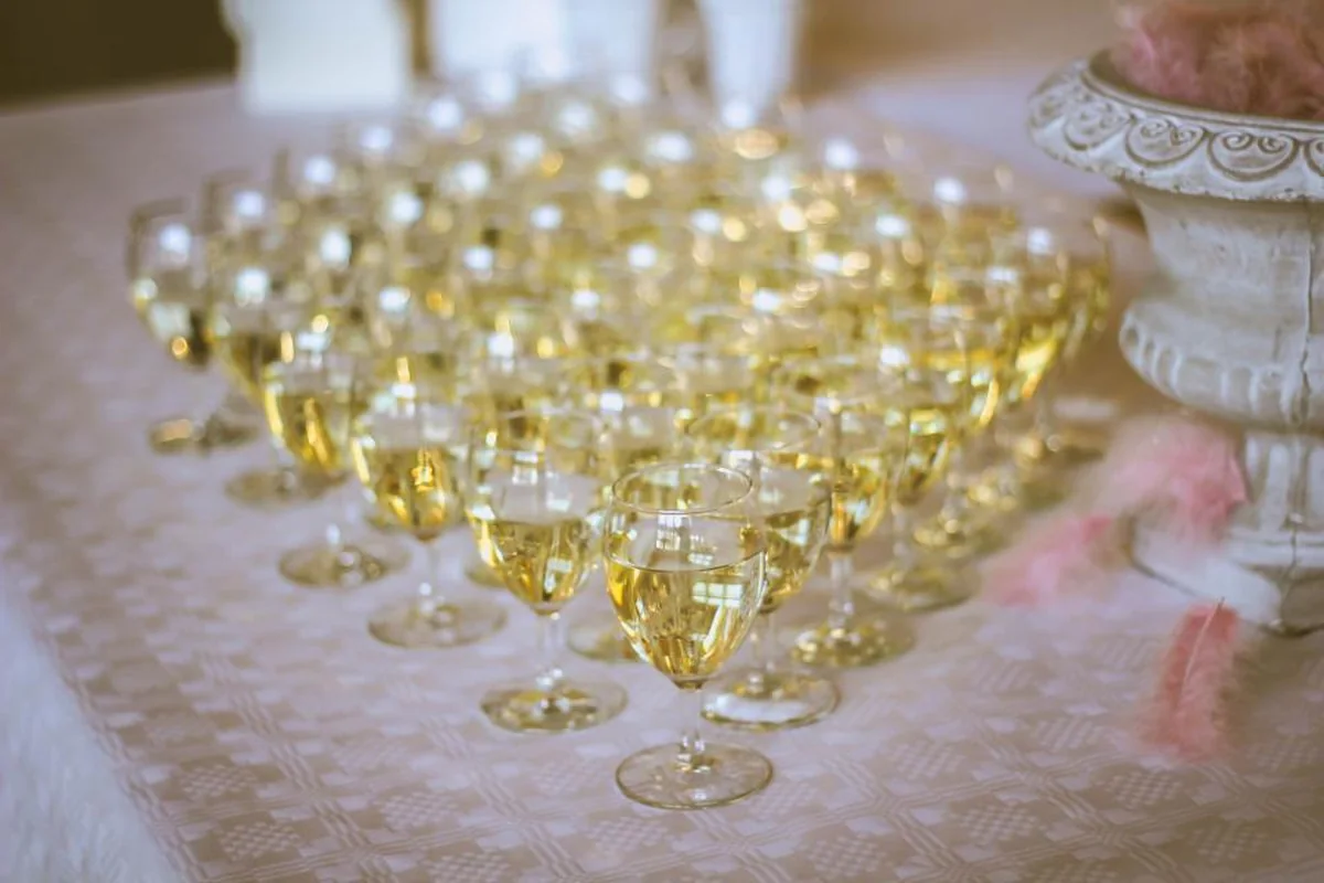 Decenas de copas de vino blanco servidas y alineadas sobre un mantel claro, junto a un jarrón de piedra con plumas rosas