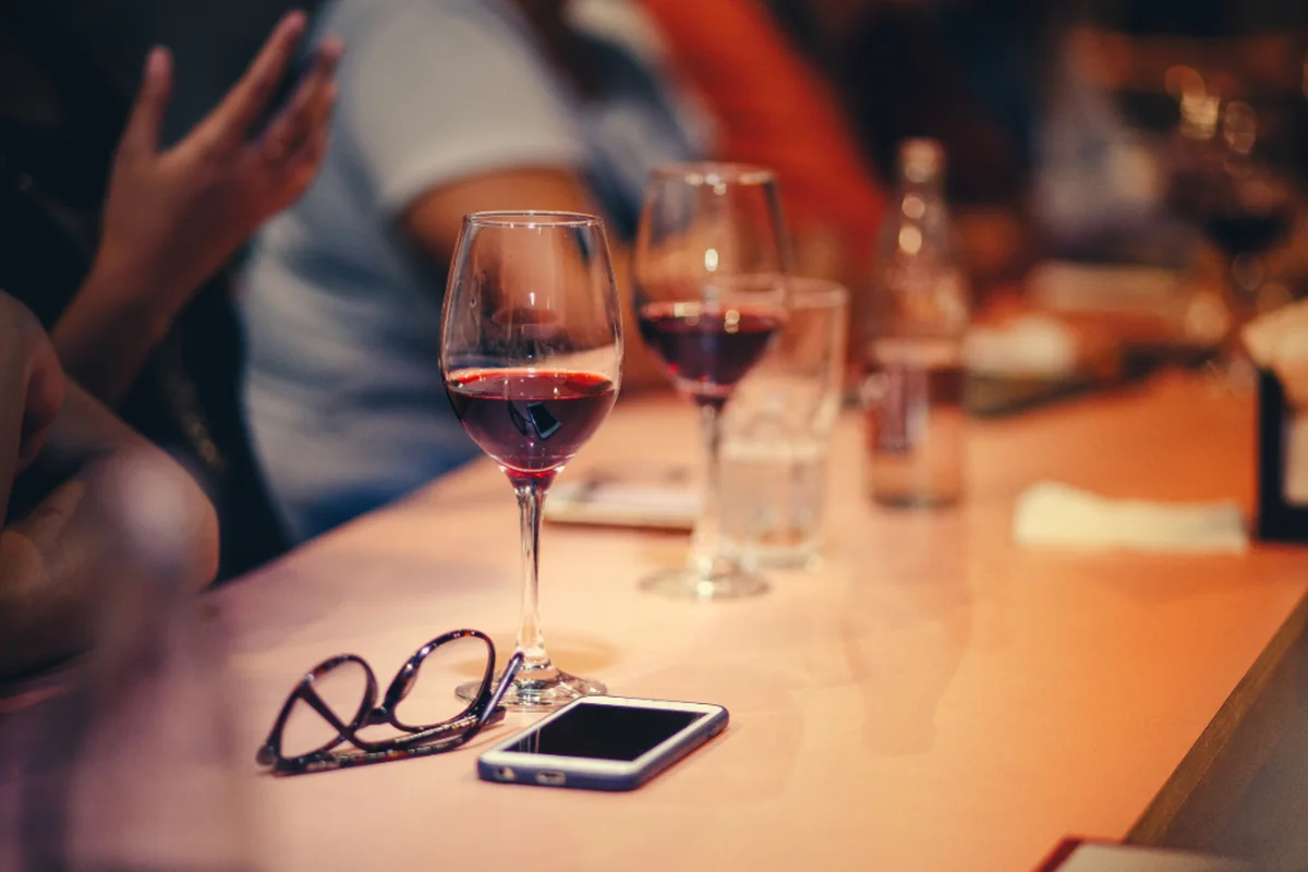 Mesa alargada de un bar con varias copas de vino tinto, un vaso de agua, unas gafas y un móvil, con varias personas desenfocadas alrededor