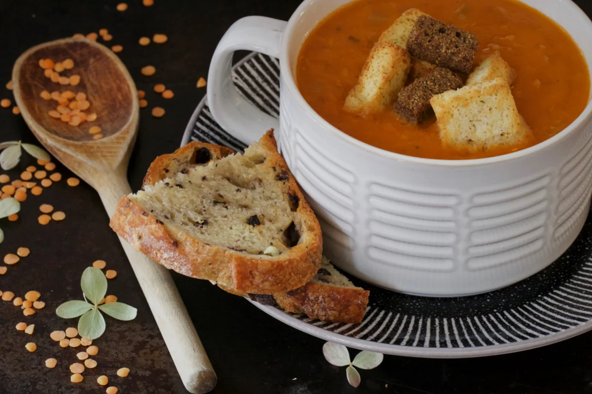 Una taza blanca con crema de lentejas y picatostes, junto a una rebanada de pan y una cuchara de madera sobre una mesa oscura