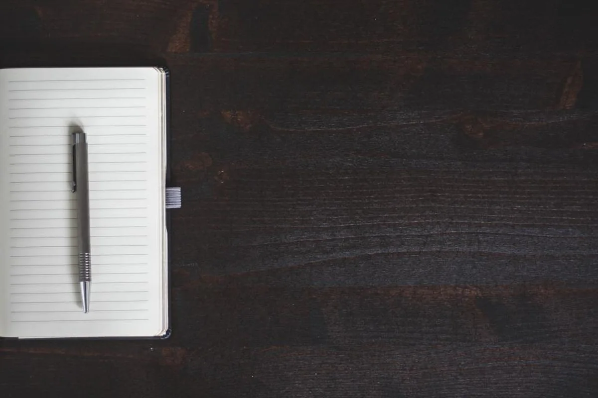 Cuaderno abierto con hojas rayadas en blanco y un bolígrafo plateado encima, sobre una mesa de madera oscura
