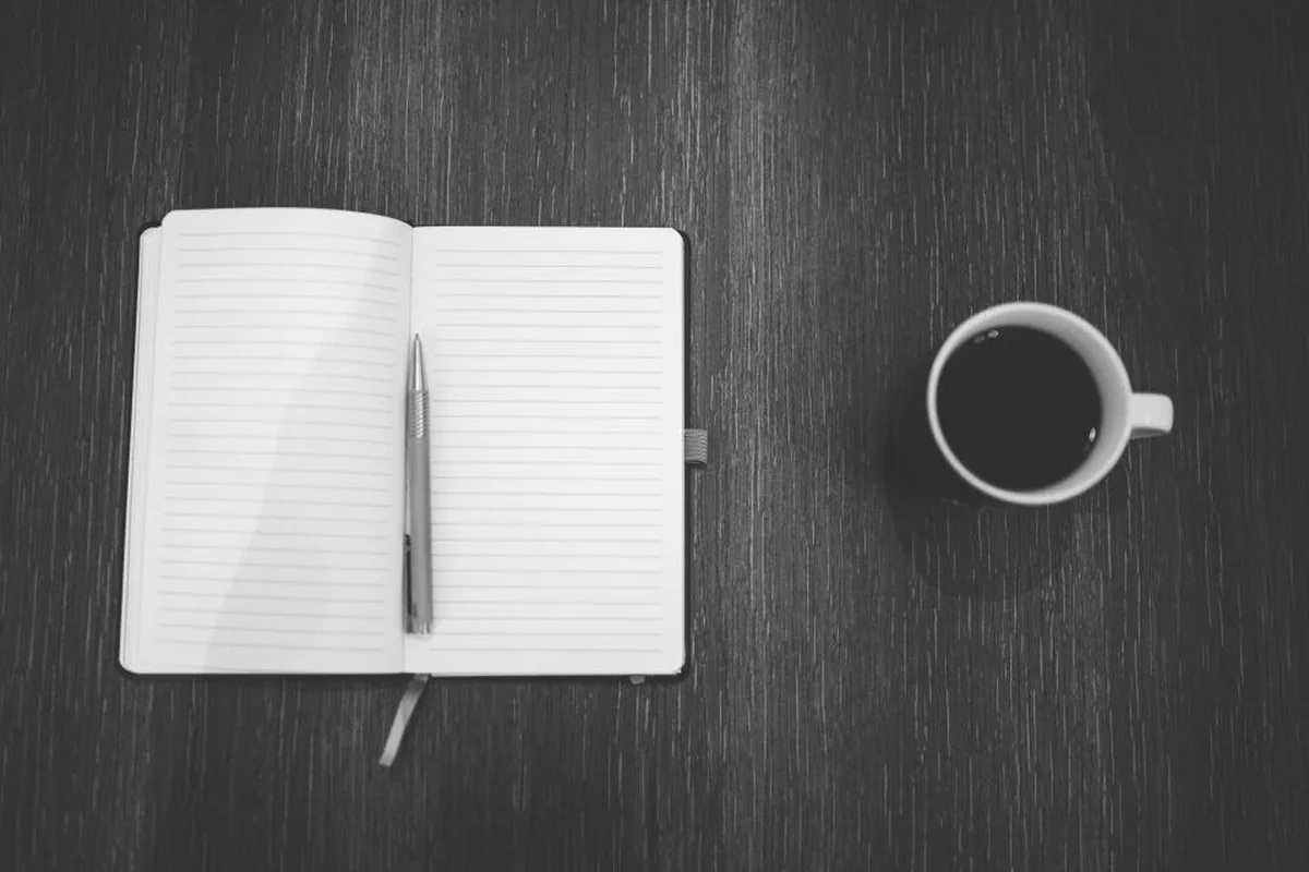 Cuaderno abierto en blanco con un bolígrafo encima y una taza de café sobre una mesa oscura